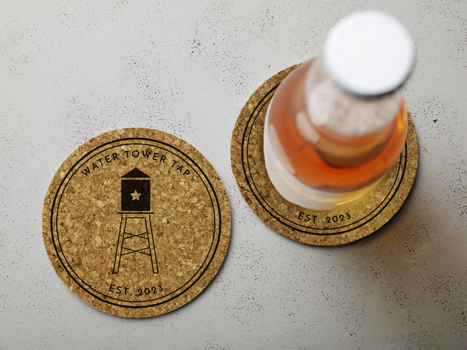 Two coasters sit on white surface with the words Water Tower Tap and an image of a water tower printed on it. One coaster has a drink on it.