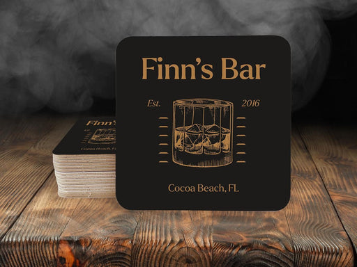 Single coaster sits on in front of a stack of coasters. Coasters feature bar design. This design uses yellow lettering on a dark brown background, with a drawing of a whiskey glass. The name, established date, and location can be customized.