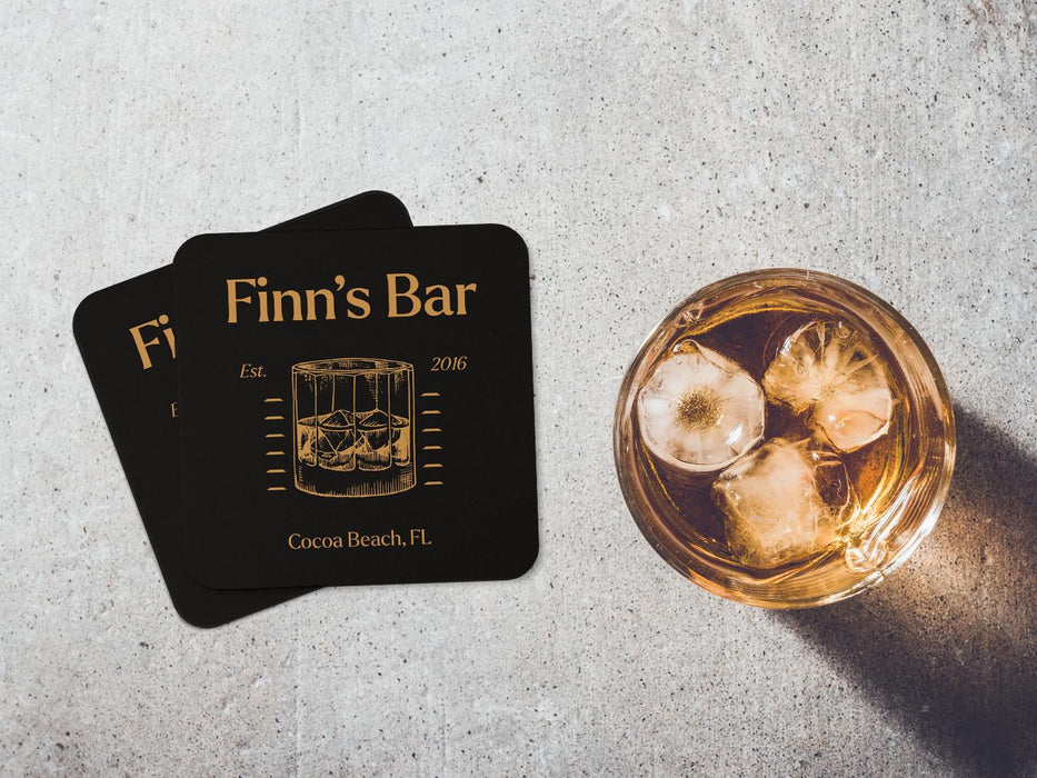 Two coasters sit on concrete background next to whiskey glass. Coasters feature bar design. This design uses yellow lettering on a dark brown background, with a drawing of a whiskey glass. The name, established date, and location can be customized.