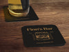 Three coasters sit on wood surface, one has a beer glass on it. Coasters feature bar design. This design uses yellow lettering on a dark brown background, with a drawing of a whiskey glass. The name, established date, and location can be customized.