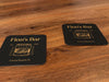 Two coasters sit on wood background. Coasters feature bar design. This design uses yellow lettering on a dark brown background, with a drawing of a whiskey glass. The name, established date, and location can be customized.