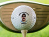 A golf ball is shown sitting on a golf tee with a golf club and green grass behind it. Ball features custom printed text and photo.