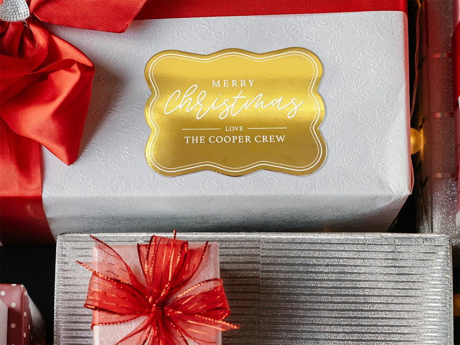 A gold foil cardstock gift tag is shown on a Christmas present. The present is wrapped in white paper and has a red bow and ribbon around it. It has other Christmas presents surrounding it.