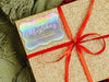 A iridescent foil cardstock gift tag is shown on a glitter wrapped box. The box also has a red ribbon on it. The box is seen on a green blanket surface.