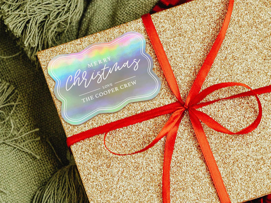 A iridescent foil cardstock gift tag is shown on a glitter wrapped box. The box also has a red ribbon on it. The box is seen on a green blanket surface.