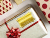 A gold foil cardstock gift tag is shown on a Christmas present. The present is wrapped in gold paper and has a red bow and ribbon around it. The background of the picture is a white surface.