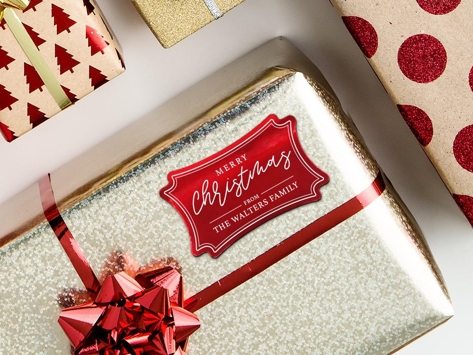 A red foil cardstock gift tag is shown on a Christmas present. The present is wrapped in gold paper and has a red bow and ribbon around it. The background of the picture is a white surface.