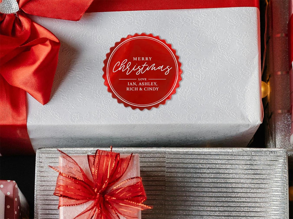 A red foil cardstock gift tag is shown on a Christmas present. The present is wrapped in white paper and has a red bow and ribbon around it. It has other Christmas presents surrounding it.