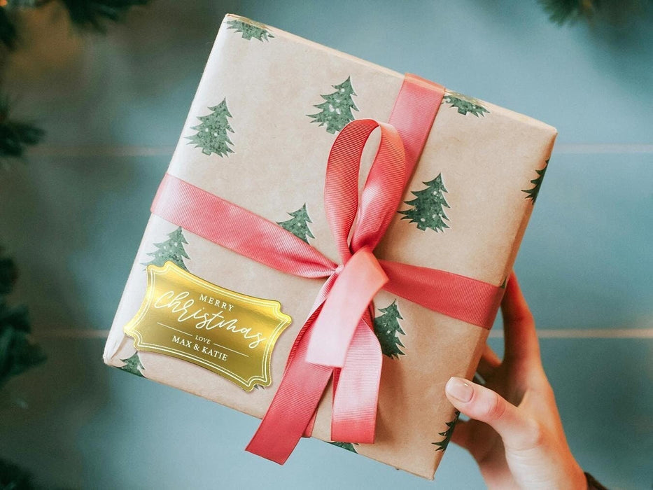 A gold foil cardstock gift tag is shown on a Christmas present. The present is wrapped in Christmas tree paper and has a red ribbon around it. The background of the picture is a green/blue.