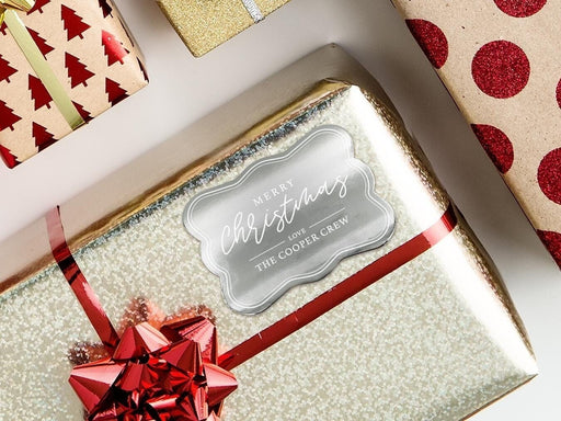 A silver foil cardstock gift tag is shown on a Christmas present. The present is wrapped in gold paper and has a red bow and ribbon around it. The background of the picture is a white surface.