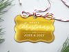 A gold foil cardstock Merry Christmas gift tag is shown sitting on a white background surrounded by Christmas tree clippings. The tag has a red and white string looped through the top of it.