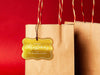 A gold foil cardstock Merry Christmas gift tag is shown hanging from a brown, craft paper gift bag. The bag is on a red backdrop.