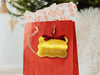 A gold foil cardstock Merry Christmas gift tag is shown hanging from red gift bag. The bag sits under a Christmas tree against a white background.