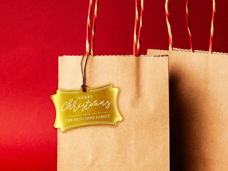 A gold foil cardstock Merry Christmas gift tag is shown hanging from a brown, craft paper gift bag. The bag is on a red backdrop.