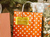 A gold foil cardstock Merry Christmas gift tag is shown hanging from a star-patterned white and red gift bag. The bag sits under a Christmas tree surrounded by other presents.