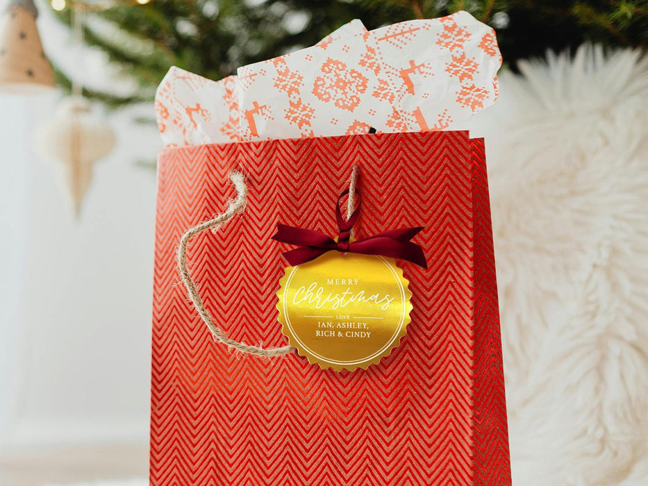 A gold foil cardstock Merry Christmas gift tag is shown hanging from red gift bag. The bag sits under a Christmas tree against a white background.