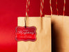 A red foil cardstock Merry Christmas gift tag is shown hanging from a brown, craft paper gift bag. The bag is on a red backdrop.