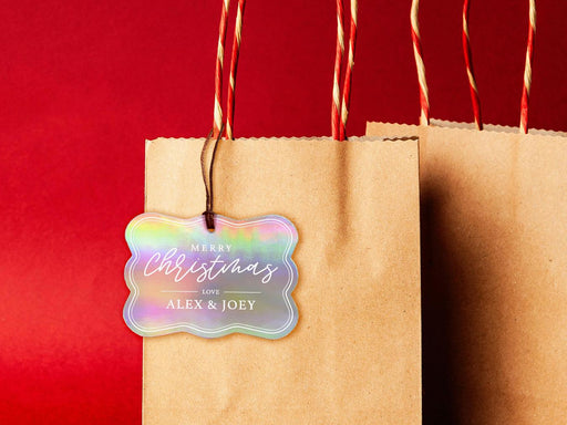 A iridescent cardstock Merry Christmas gift tag is shown hanging from a brown, craft paper gift bag. The bag is on a red backdrop.