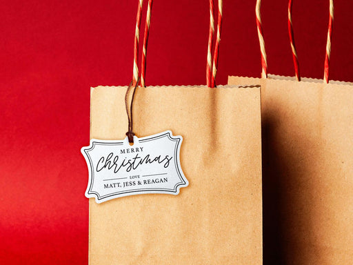 A white glitter cardstock Merry Christmas gift tag with black ink is shown hanging from a brown, craft paper gift bag. The bag is on a red backdrop.