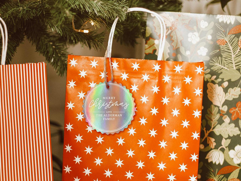 An iridescent cardstock Merry Christmas gift tag is shown hanging from a star-patterned white and red gift bag. The bag sits under a Christmas tree surrounded by other presents.