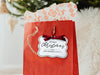 A white glitter cardstock Merry Christmas gift tag with black ink is shown hanging from red gift bag. The bag sits under a Christmas tree against a white background.