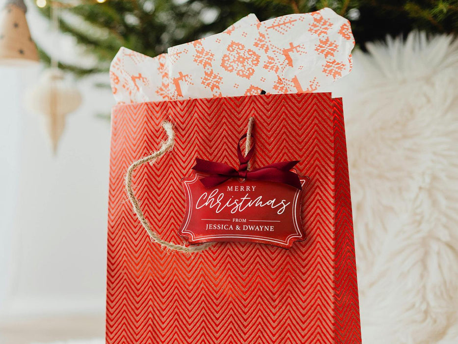 A red foil cardstock Merry Christmas gift tag is shown hanging from red gift bag. The bag sits under a Christmas tree against a white background.