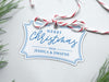 A white glitter cardstock Merry Christmas gift tag with blue ink is shown sitting on a white background surrounded by Christmas tree clippings. The tag has a red and white string looped through the top of it.