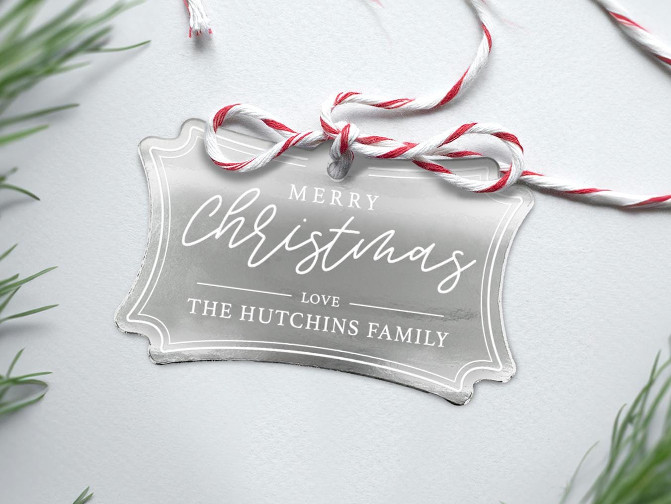 A silver foil cardstock Merry Christmas gift tag is shown sitting on a white background surrounded by Christmas tree clippings. The tag has a red and white string looped through the top of it.