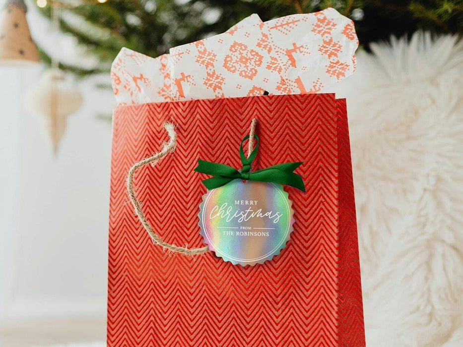 An iridescent cardstock Merry Christmas gift tag is shown hanging from red gift bag. The bag sits under a Christmas tree against a white background.