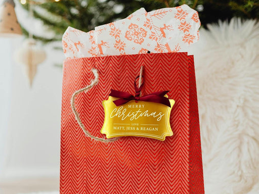 A gold foil cardstock Merry Christmas gift tag is shown hanging from red gift bag. The bag sits under a Christmas tree against a white background.