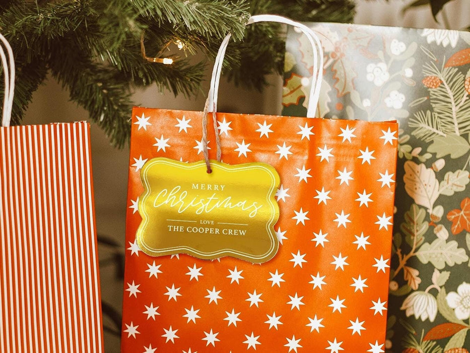 A gold foil cardstock Merry Christmas gift tag is shown hanging from a star-patterned white and red gift bag. The bag sits under a Christmas tree surrounded by other presents.