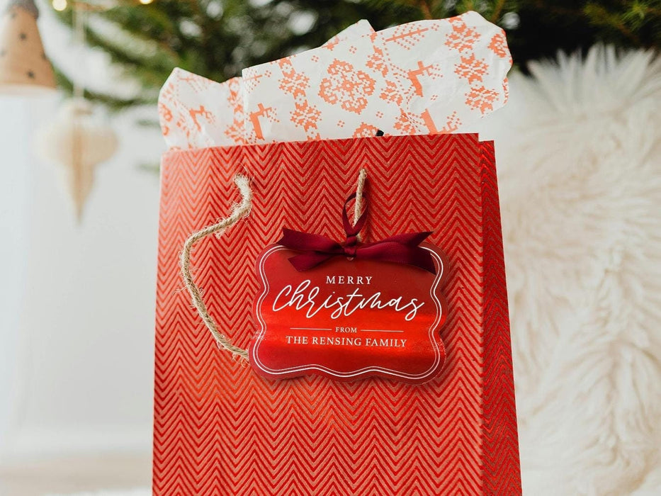 A red foil cardstock Merry Christmas gift tag is shown hanging from red gift bag. The bag sits under a Christmas tree against a white background.