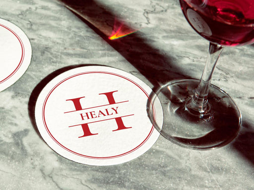 Two red monogram coasters are shown on a marble surface with a wine glass on top. The coasters display the name Healy with a lined circular frame around it.