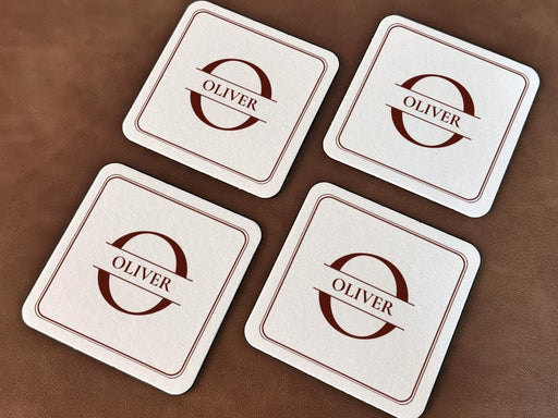 A stack of brown monogram coasters are shown on a leather surface. The coasters display the name Oliver with a lined square frame around it.