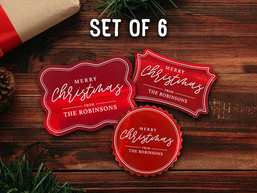 Text reads, Set of 6. Three red foil cardstock Merry Christmas gift tags are shown on a dark wooden surface. Pine tree branches and a Christmas present are shown around the tags.