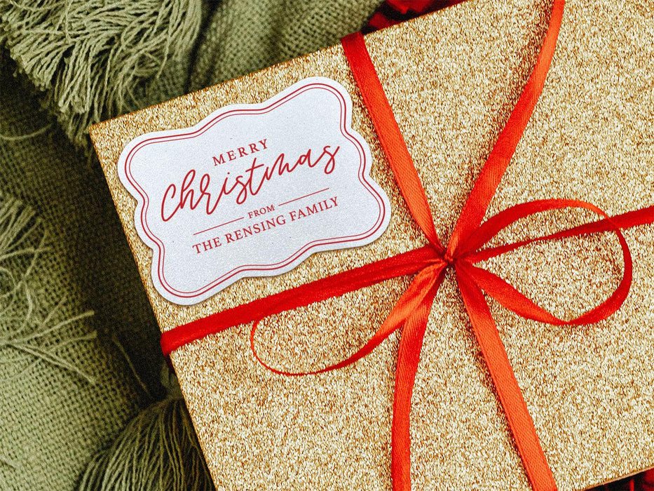 A white glitter cardstock gift tag printed with red ink is shown on a glitter wrapped box. The box also has a red ribbon on it. The box is seen on a green blanket surface.