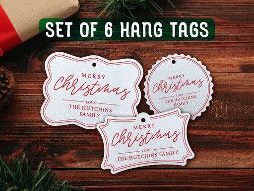 Text reads, Set of 6. Three white glitter cardstock Merry Christmas hanging gift tags with red ink are shown on a dark wooden surface. Pine tree branches, a pine cone, a red ornament, and a Christmas present are shown around the tags.