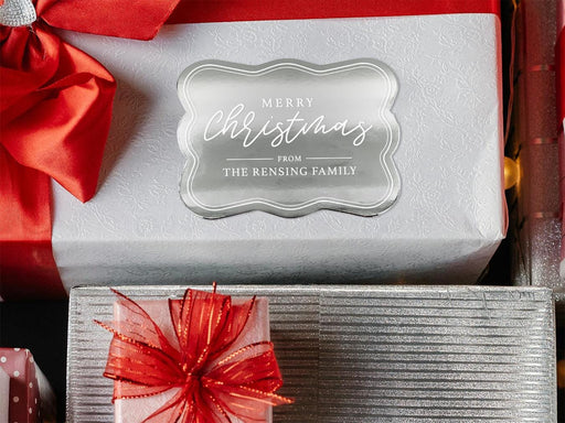 A silver foil cardstock gift tag is shown on a Christmas present. The present is wrapped in white paper and has a red bow and ribbon around it. It has other Christmas presents surrounding it.