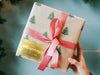 A gold foil cardstock gift tag is shown on a Christmas present. The present is wrapped in Christmas tree paper and has a red ribbon around it. The background of the picture is a green/blue.