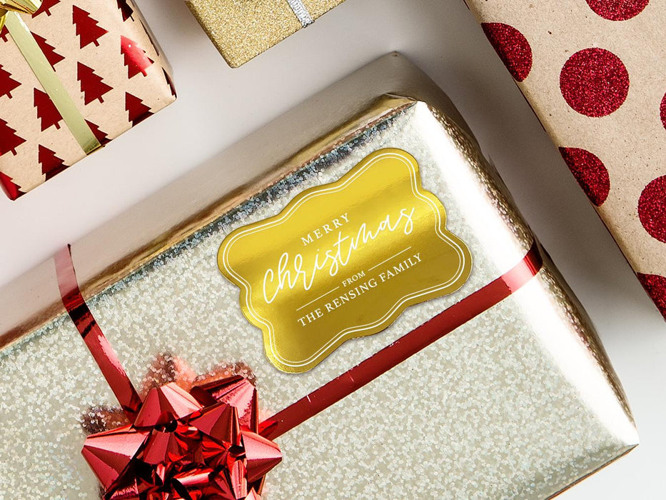 A gold foil cardstock gift tag is shown on a Christmas present. The present is wrapped in gold paper and has a red bow and ribbon around it. The background of the picture is a white surface.