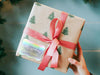 A iridescent foil cardstock gift tag is shown on a Christmas present. The present is wrapped in Christmas tree paper and has a red ribbon around it. The background of the picture is a green/blue.