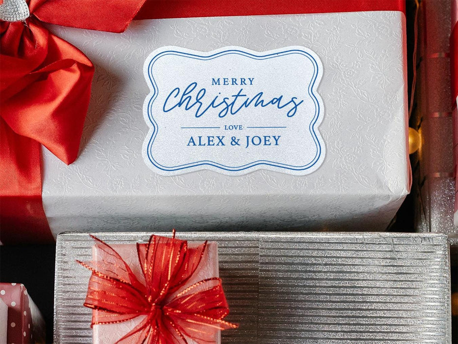 A white glitter cardstock gift tag printed with blue ink is shown on a Christmas present. The present is wrapped in white paper and has a red bow and ribbon around it. It has other Christmas presents surrounding it.