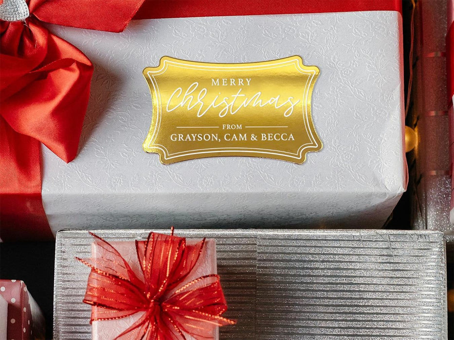 A gold foil cardstock gift tag is shown on a Christmas present. The present is wrapped in white paper and has a red bow and ribbon around it. It has other Christmas presents surrounding it.