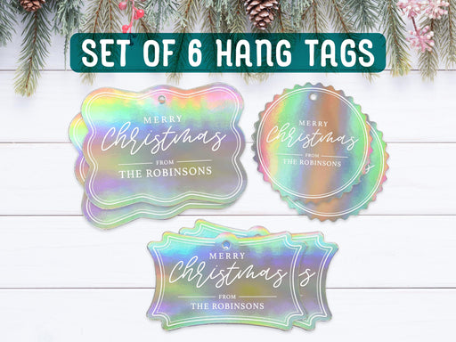 Text reads, Set of 6 Hang Tags. Six iridescent foil cardstock Merry Christmas hang tags are shown on a dark wooden surface. Pine tree branches, gold ornaments, and a Christmas present are shown around the tags.