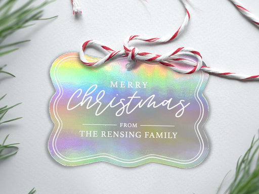 An iridescent foil cardstock Merry Christmas gift tag is shown sitting on a white background surrounded by Christmas tree clippings. The tag has a red and white string looped through the top of it.