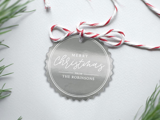 A silver foil cardstock Merry Christmas gift tag is shown sitting on a white background surrounded by Christmas tree clippings. The tag has a red and white string looped through the top of it.