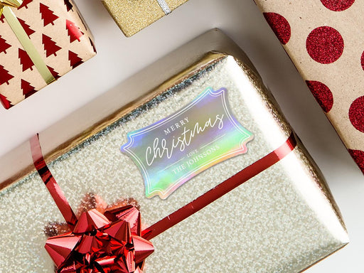 An iridescent foil cardstock Merry Christmas gift tag is shown on a glitter wrapped box. The box also has a red ribbon on it. The box is seen on a green blanket surface.