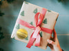 A gold foil cardstock gift tag is shown on a Christmas present. The present is wrapped in Christmas tree paper and has a red ribbon around it. The background of the picture is a green/blue.