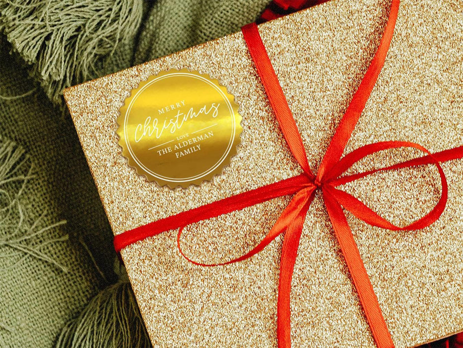 A gold foil cardstock gift tag is shown on a glitter wrapped box. The box also has a red ribbon on it. The box is seen on a green blanket surface.