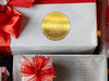 A gold foil cardstock gift tag is shown on a Christmas present. The present is wrapped in white paper and has a red bow and ribbon around it. It has other Christmas presents surrounding it.